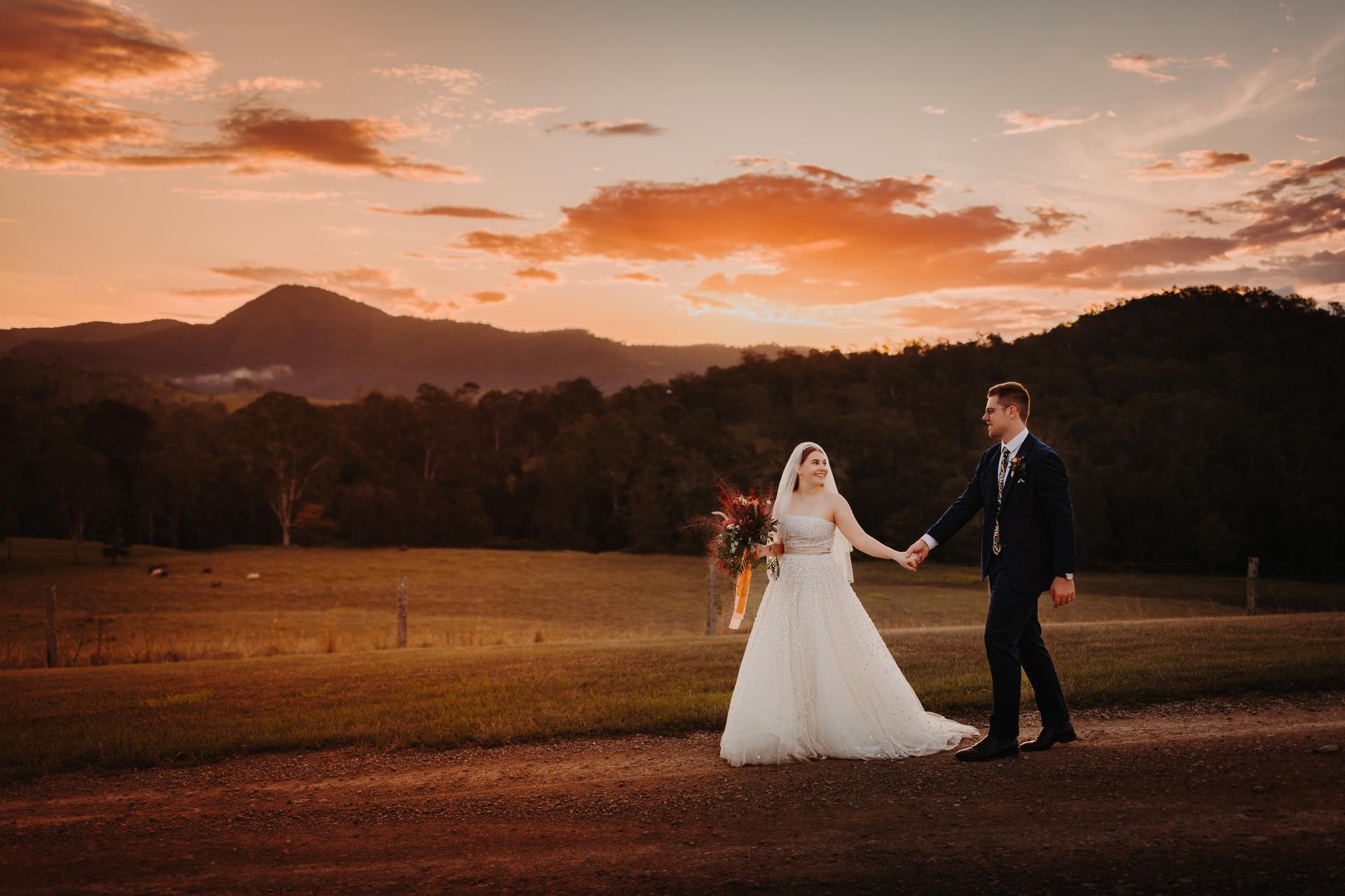 Bride Lilly and groom Connor hold hands and walk on a dirt path at Yabbaloumba Retreat during sunset, with hills and trees in the background.