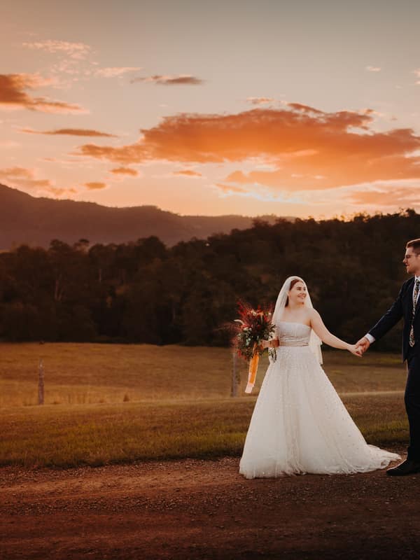Bride Lilly and groom Connor hold hands and walk on a dirt path at Yabbaloumba Retreat during sunset, with hills and trees in the background.