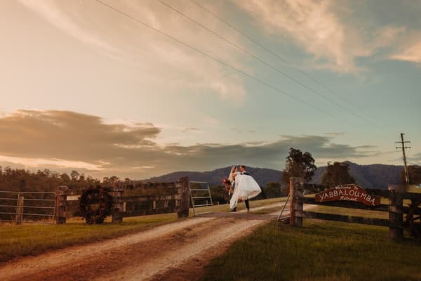 The groom carries the bride on a dirt road at the entrance of Yabbaloumba Retreat with hills and trees in the background under a partly cloudy sky at sunset.