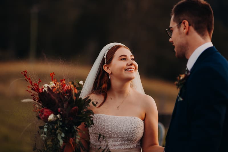 The bride Lilly in a strapless white wedding dress and veil holding a bouquet of flowers looks smilingly at the groom Connor, who is wearing a dark suit and glasses, at Yabbaloumba Retreat.