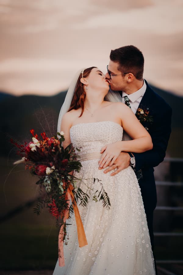 Bride Lilly and groom Connor share a kiss during their couple portraits at Yabbaloumba Retreat. Lilly wears a strapless white wedding dress and holds a bouquet with red and green foliage, while Connor wears a dark suit with a floral boutonniere.