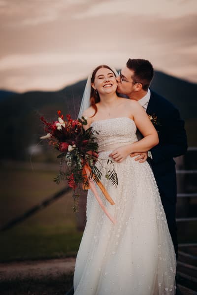 Bride Lilly in a strapless white wedding gown holding a bouquet of flowers is embraced from behind and kissed on the cheek by groom Connor in a dark suit at Yabbaloumba Retreat with hills in the background.