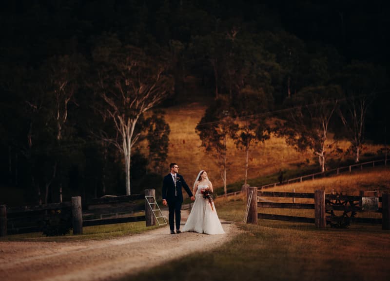 Bride Lilly and groom Connor walk hand in hand along a dirt path at Yabbaloumba Retreat, surrounded by wooden fences and trees in a natural outdoor setting.