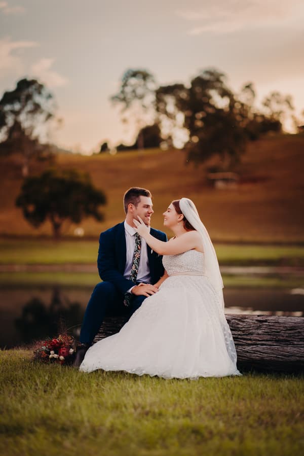 Bride Lilly and groom Connor sit on a large log near a pond at Yabbaloumba Retreat, with Lilly gently touching Connor's face during their couple portraits session.