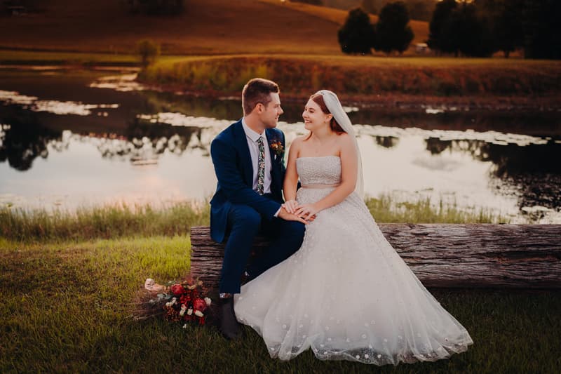 Bride Lilly and groom Connor sit on a log by a lake at Yabbaloumba Retreat, holding hands and looking at each other. Lilly wears a strapless white wedding gown with a veil, and Connor wears a blue suit with a floral tie. A bouquet lies on the grass beside them.
