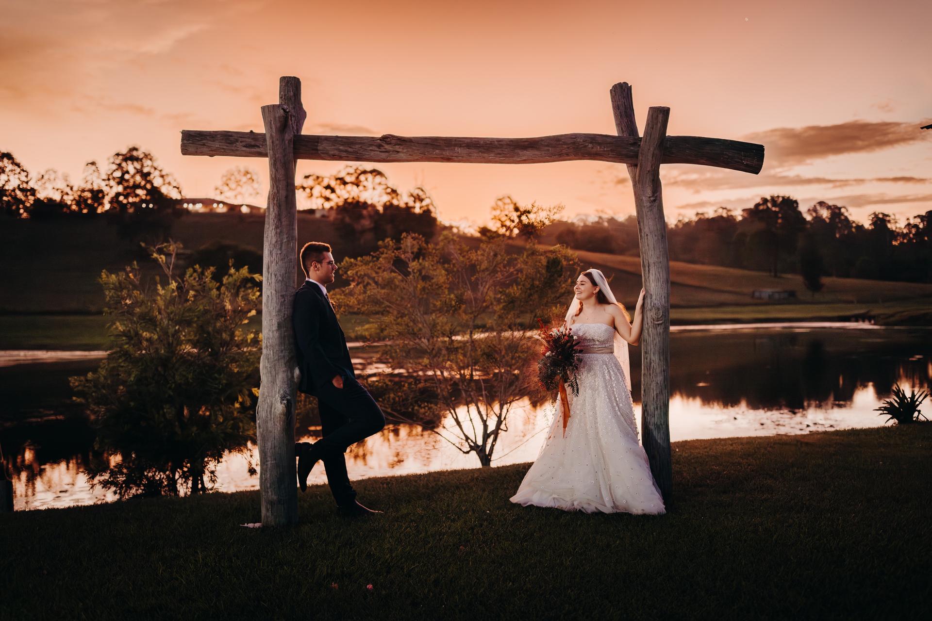 Bride Lilly and groom Connor pose under a rustic wooden arch near a lake at Yabbaloumba Retreat during sunset.