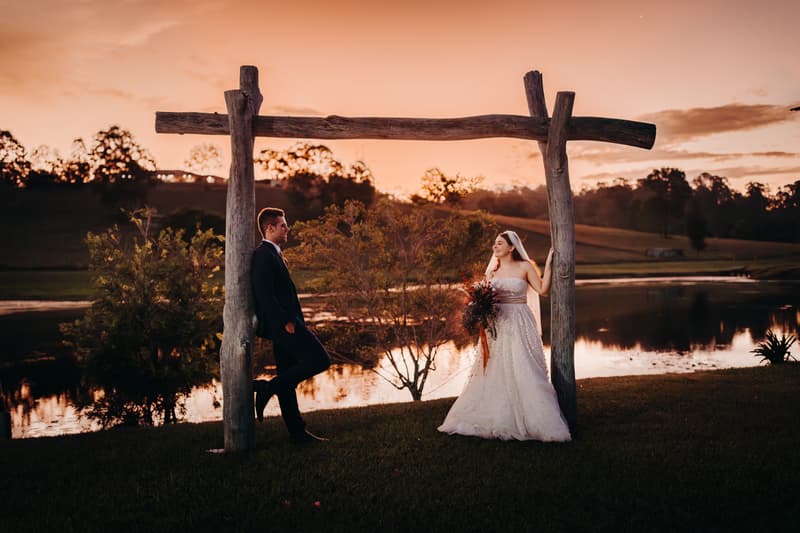Bride Lilly and groom Connor pose under a rustic wooden arch near a lake at Yabbaloumba Retreat during sunset.