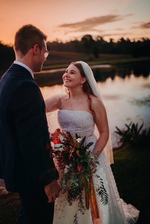 Bride Lilly and groom Connor stand facing each other near a lake at Yabbaloumba Retreat during sunset. Lilly holds a large bouquet of flowers and wears a strapless white wedding gown with a veil, while Connor wears a dark suit and glasses.