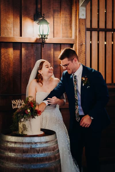 Bride Lilly and groom Connor cut their wedding cake together, standing beside a wooden barrel with a floral-decorated cake on top. Valentine's Day wedding photography by She Said Yes.