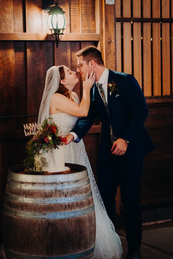 Bride Lilly and groom Connor lean in for a kiss while cutting their wedding cake on a wooden barrel at Yabbaloumba Retreat — The Shed.