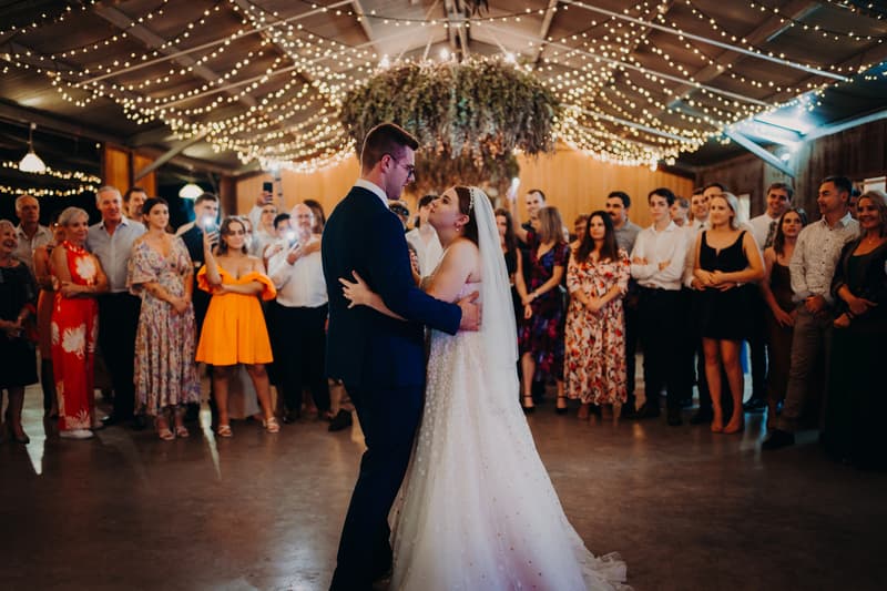 Bride Lilly and groom Connor share a dance at the reception in The Shed at Yabbaloumba Retreat, surrounded by guests watching and taking photos under string lights and hanging greenery.