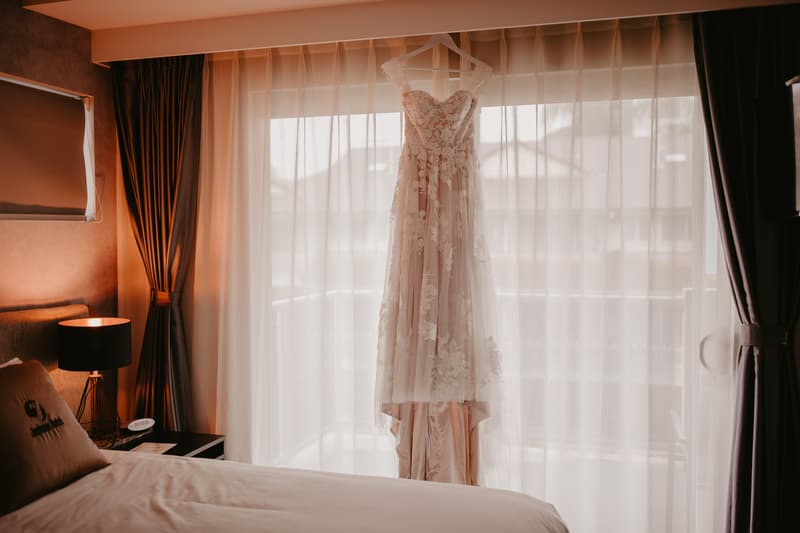 A wedding dress with lace details hangs on a hanger in front of sheer curtains covering a window in a softly lit hotel room.