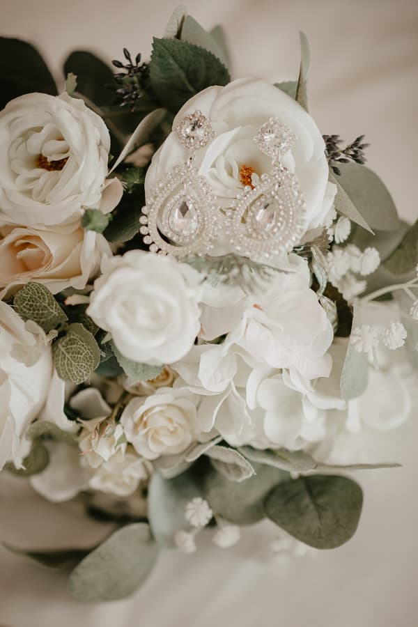 Close-up of a bridal bouquet featuring white and cream flowers with green leaves, adorned with a pair of ornate crystal and pearl earrings placed on top.
