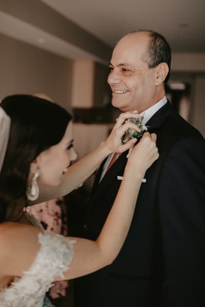 The bride adjusts the boutonniere on an older man's suit jacket indoors. Engagement celebration by She Said Yes.