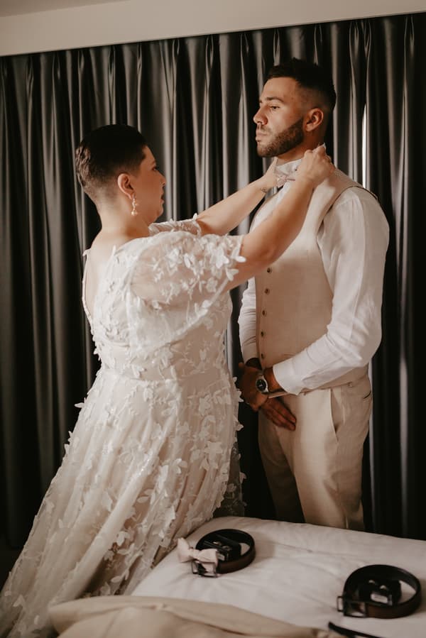 A bride adjusts the groom's bow tie as he stands facing her in a room with dark curtains.