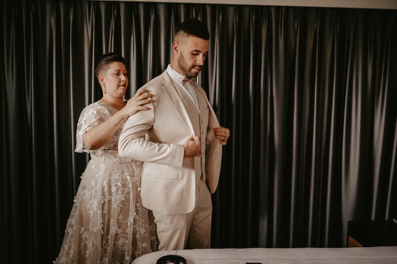 A bride adjusts the groom's beige suit jacket in front of dark curtains indoors.