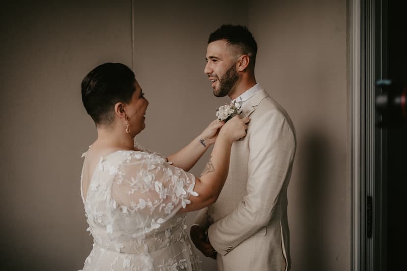 The bride adjusts the groom's boutonniere on his beige suit jacket against a plain wall background.