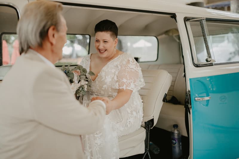 A bride in a white floral lace dress holding a bouquet sits inside a vintage blue and white van, holding hands with an older man dressed in a light-colored suit standing outside the vehicle.