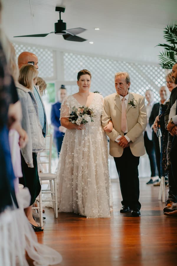 The bride in a white floral lace gown holding a bouquet walks arm-in-arm with an older man in a beige suit and pink tie down an aisle lined with seated guests.