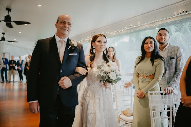 The bride, holding a bouquet, is walking arm-in-arm with an older man in a suit, while guests stand and watch in a decorated indoor venue with string lights.