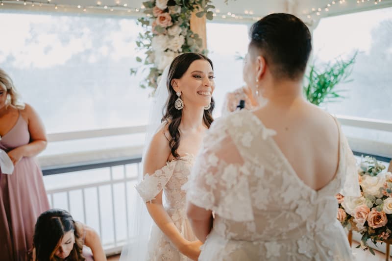 Two brides in lace wedding dresses face each other indoors near a floral arrangement and string lights, with two bridesmaids in mauve dresses in the background.