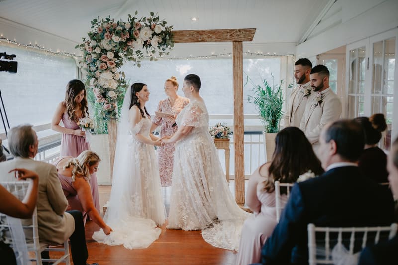Two brides stand holding hands under a floral arch during their wedding ceremony, with an officiant reading from a book behind them. Two groomsmen stand to the right, and guests are seated facing the couple. A bridesmaid adjusts the train of one bride's dress on the left.