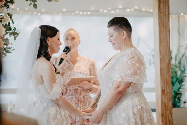 Two brides in white lace wedding dresses hold hands facing each other during a ceremony, with an officiant standing behind them holding a microphone and a tablet.