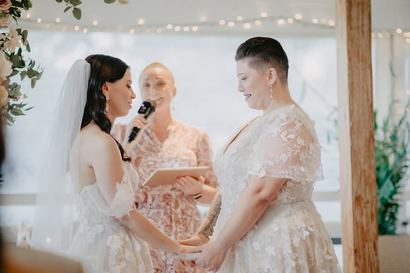 Two brides in white lace wedding dresses hold hands facing each other during a ceremony, with an officiant standing behind them holding a microphone and a tablet.