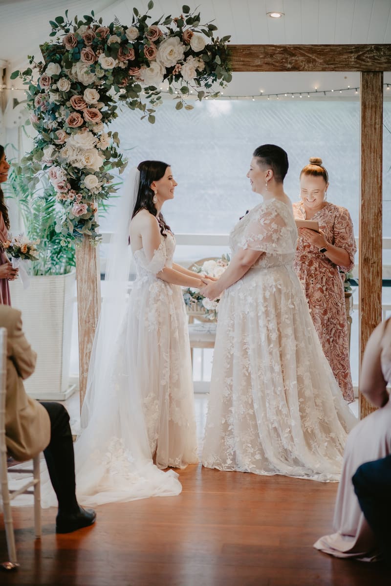 Two brides in lace wedding gowns hold hands facing each other under a wooden arch decorated with flowers, while an officiant reads from a tablet behind them during a wedding ceremony.