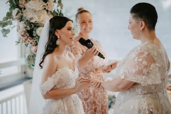 Two brides hold hands and exchange vows during their wedding ceremony, with an officiant holding a microphone and a tablet in the background.