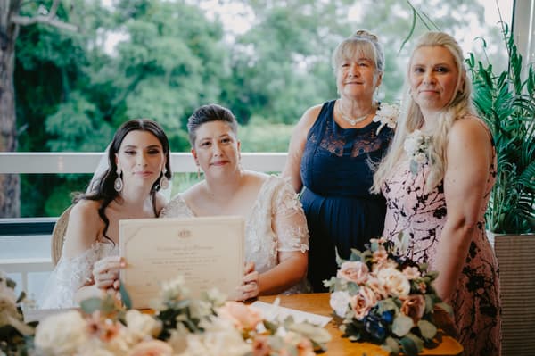 Two brides seated at a table holding a certificate of marriage, accompanied by two women standing behind them, with floral arrangements on the table and greenery visible outside the window.