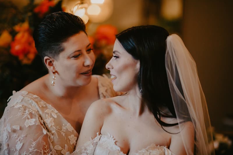 Two women in wedding attire face each other closely; one wears a lace dress with floral patterns and earrings, the other wears a strapless lace wedding gown and a veil.