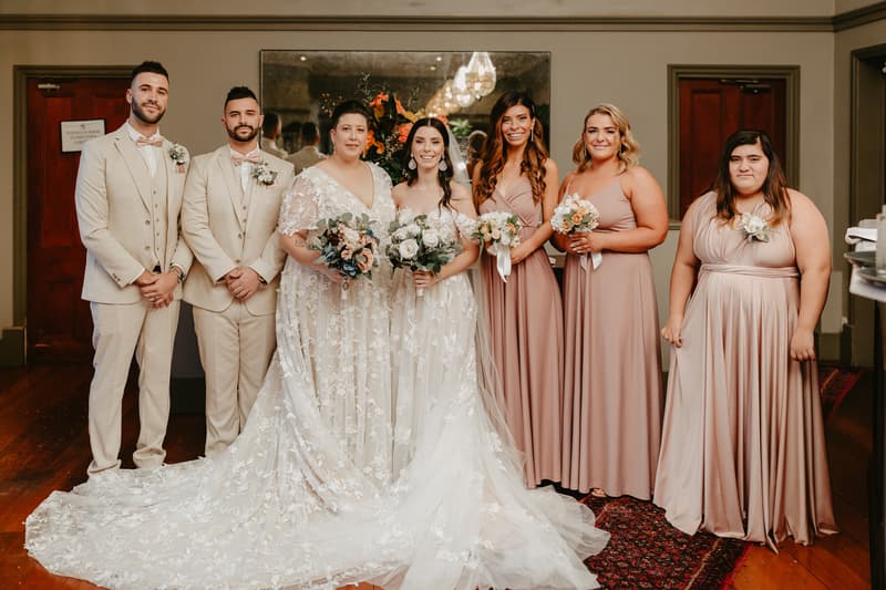A wedding party group portrait featuring two groomsmen in beige suits, the bride in a white lace gown holding a bouquet, the mother of the bride in a white lace dress holding a bouquet, and four bridesmaids in matching mauve dresses holding bouquets, standing indoors on a wooden floor with a large mirror and floral arrangement behind them.
