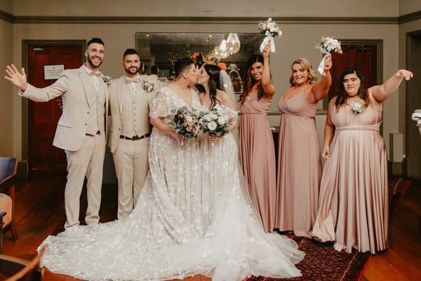 Two brides in white wedding gowns kiss while holding bouquets, flanked by two groomsmen in beige suits on the left and three bridesmaids in mauve dresses holding bouquets on the right, standing indoors on a wooden floor with a large mirror behind them.