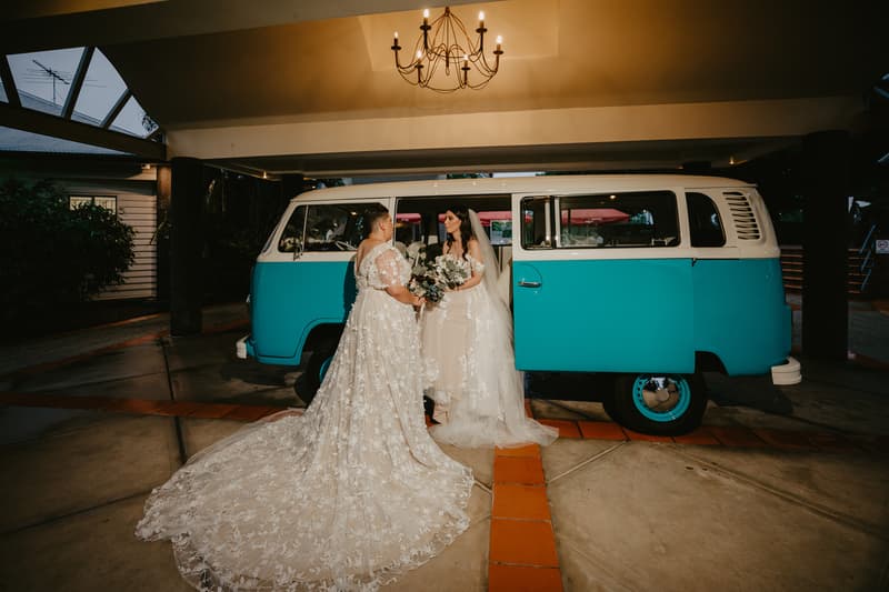 Two brides in wedding gowns stand facing each other beside a turquoise and white vintage van with the side door open under a chandelier-lit covered area.