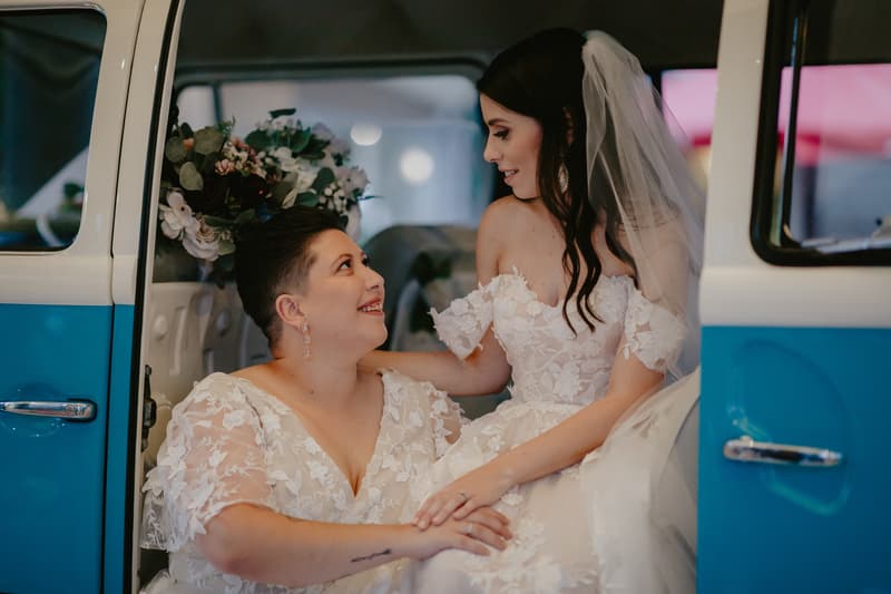 Two brides in white lace wedding dresses sit inside a blue and white vintage vehicle, holding hands and looking at each other.