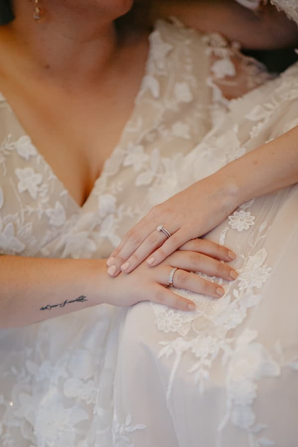 Close-up of two brides wearing white lace wedding dresses, showing their hands with wedding rings and a tattoo on one wrist.