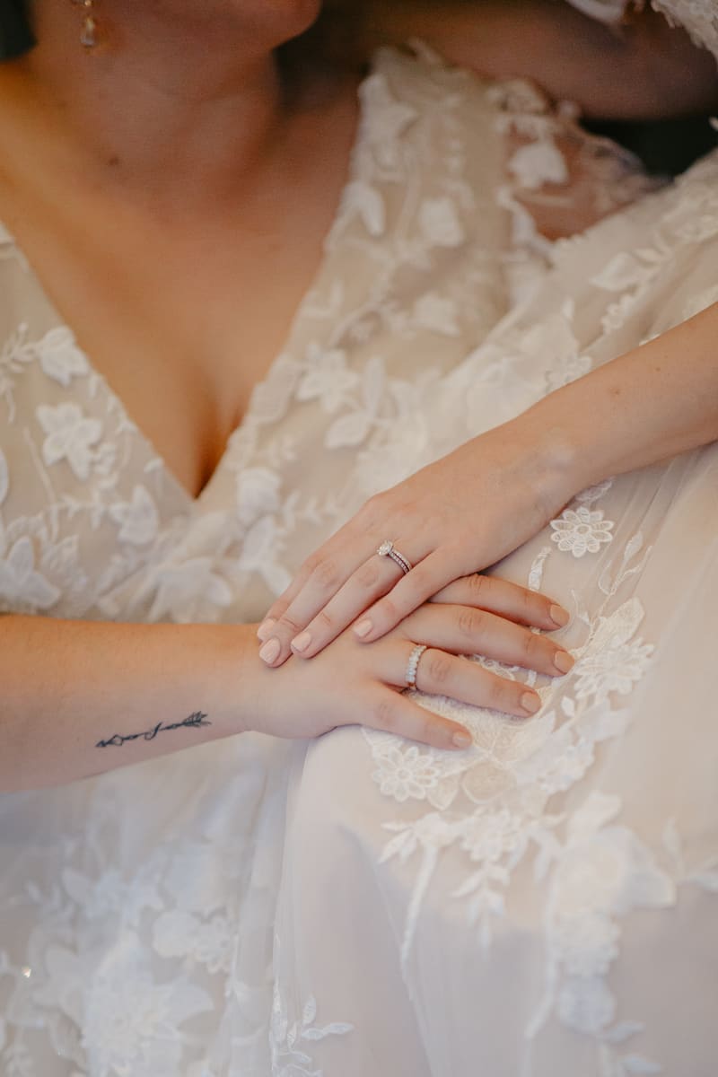 Close-up of two brides wearing white lace wedding dresses, showing their hands with wedding rings and a tattoo on one wrist. Elopement photography packages Queensland by She Said Yes.