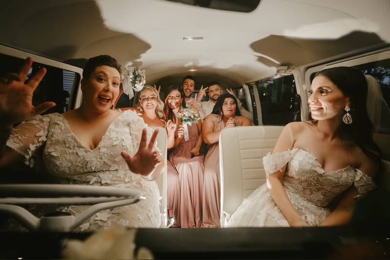 Two brides sit in the front seats of a vehicle, with bridesmaids and groomsmen seated behind them, some holding bouquets and making playful gestures.