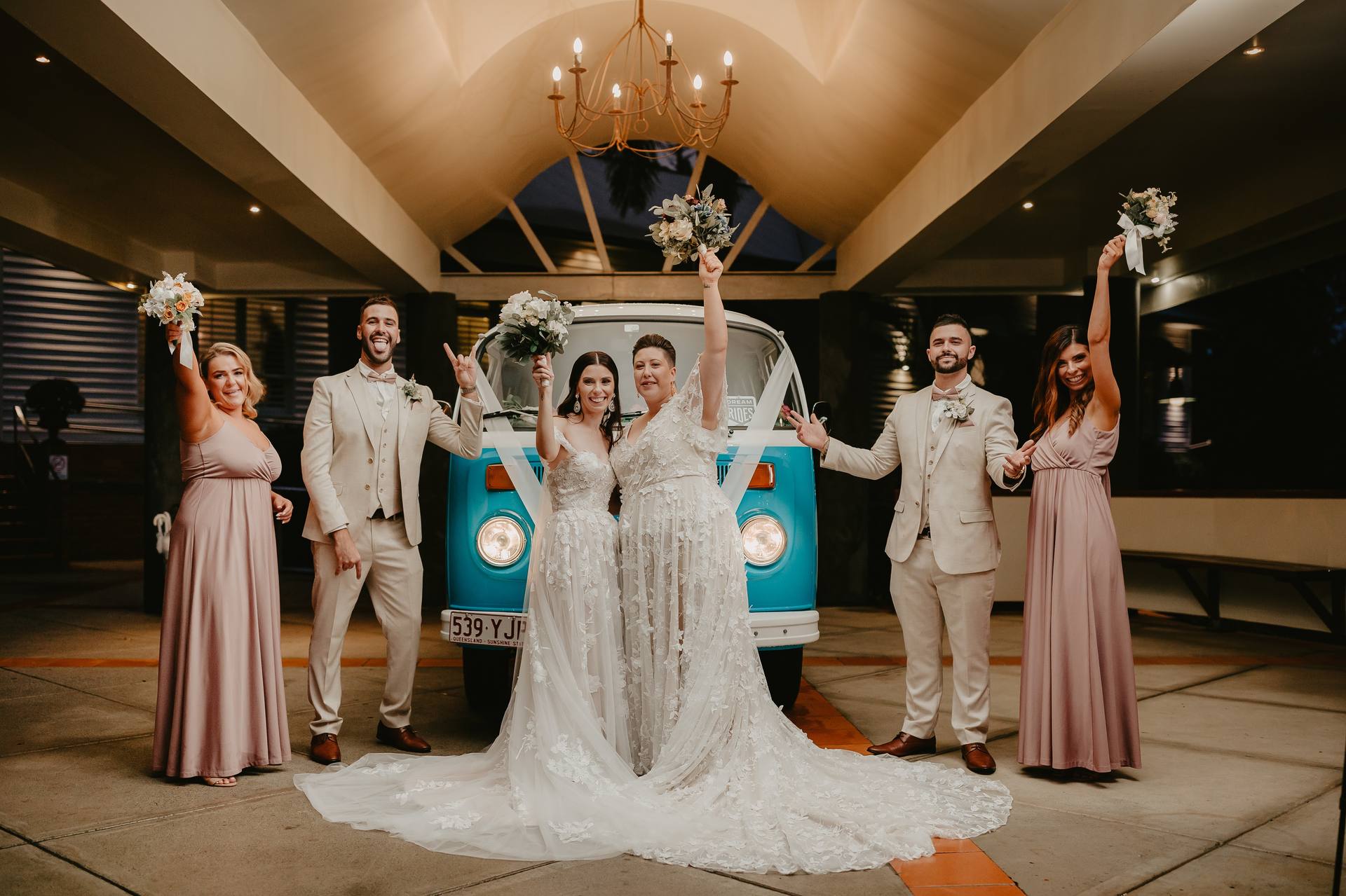 Two brides in wedding gowns stand in front of a blue vintage van with their bridal party, consisting of two bridesmaids in mauve dresses and two groomsmen in beige suits, all raising bouquets or hands in celebration under a chandelier in a covered outdoor area.