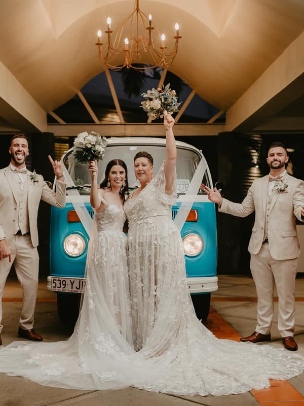 Two brides in wedding gowns stand in front of a blue vintage van with their bridal party, consisting of two bridesmaids in mauve dresses and two groomsmen in beige suits, all raising bouquets or hands in celebration under a chandelier in a covered outdoor area.