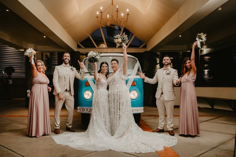 Two brides in wedding gowns stand in front of a blue vintage van with their bridal party, consisting of two bridesmaids in mauve dresses and two groomsmen in beige suits, all raising bouquets or hands in celebration under a chandelier in a covered outdoor area.