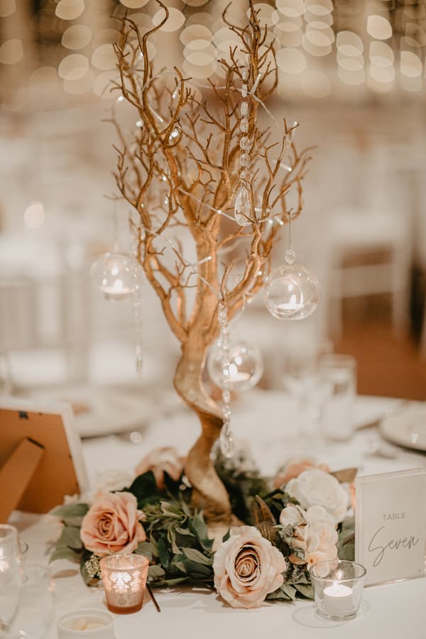 A wedding table centerpiece featuring a decorative gold tree adorned with hanging glass candle holders and crystal beads, surrounded by a floral arrangement of blush and white roses and greenery, with lit votive candles and a table number card labeled 'Table Seven'.