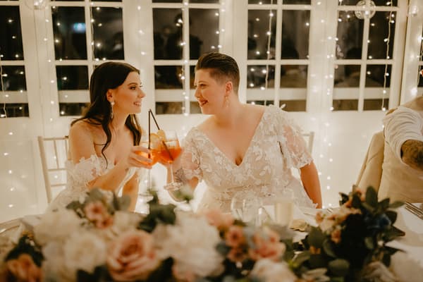 Two brides in white lace dresses sit at a decorated table with floral arrangements, clinking glasses with orange-colored drinks in front of a window with string lights.
