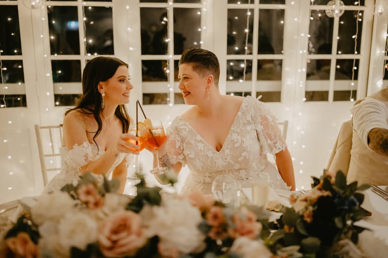 Two brides in white lace dresses sit at a decorated table with floral arrangements, clinking glasses with orange-colored drinks in front of a window with string lights.