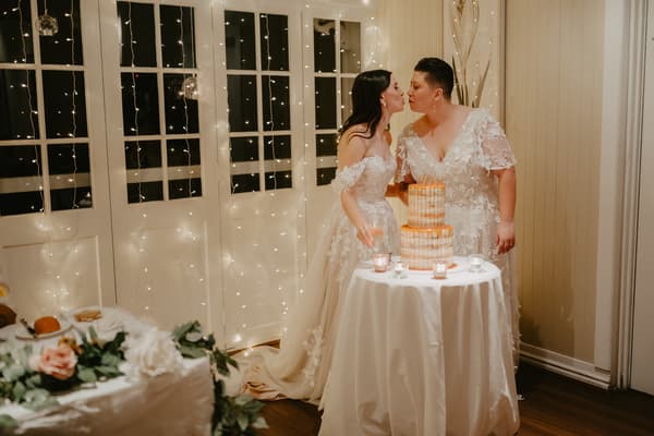 Two brides in white lace wedding dresses lean toward each other near a round table with a two-tiered cake and small candles, standing in front of a wall with string lights.
