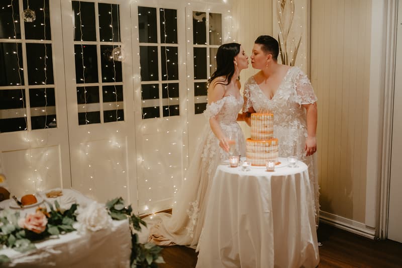 Two brides in white lace wedding dresses lean toward each other near a round table with a two-tiered cake and small candles, standing in front of a wall with string lights.