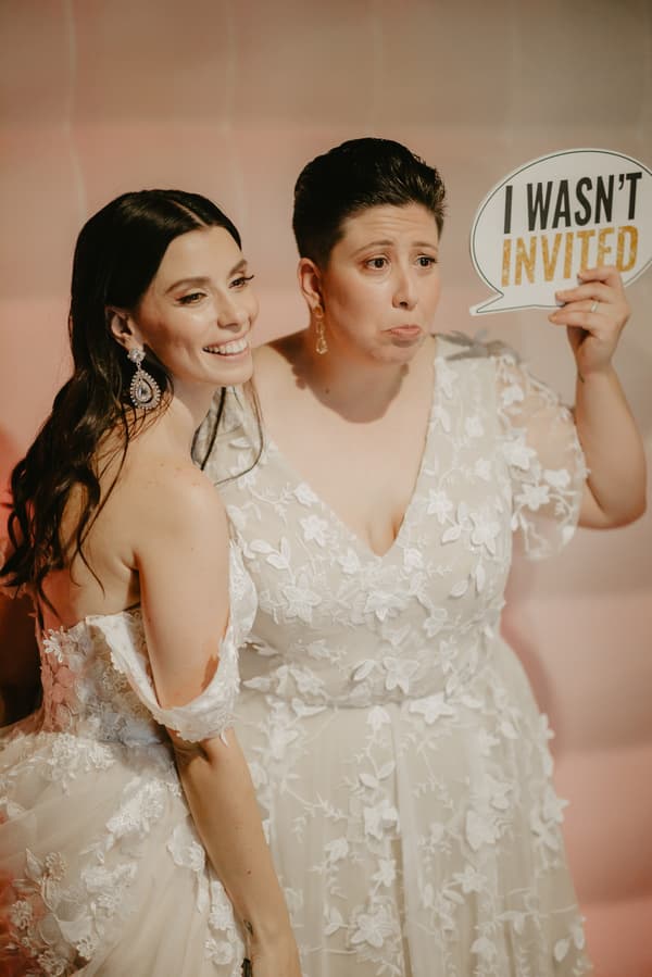Two brides in white floral lace wedding dresses pose together; one bride smiles while the other holds a speech bubble sign reading 'I WASN'T INVITED'.