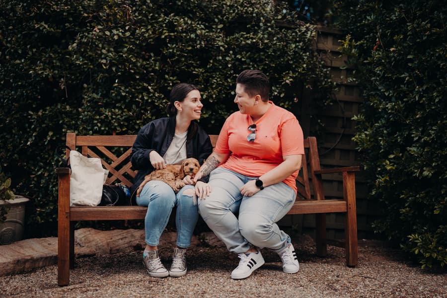The couple sitting on a wooden bench at Hillstone St Lucia, smiling and holding a small dog. The setting is lush and green.