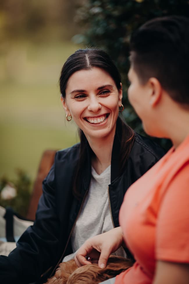 Close-up of one person smiling warmly at the other, with greenery blurred in the background.